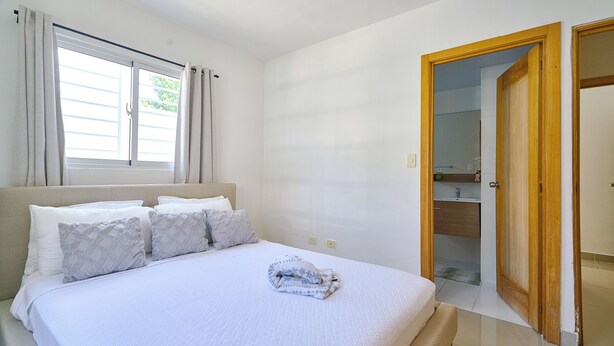 A bedroom with a bed, a window with curtains, a bathroom visible through an open door, and a tiled floor. - Charming 3-bedroom apartment with Wi-Fi, AC in welcoming Santo Domingo Este. (Santo Domingo Este)