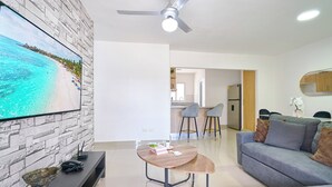 A modern living room with a flat-screen TV mounted on a textured wall, a ceiling fan, a grey sofa, and a small round table with a decorative item. - Charming 3-bedroom apartment with Wi-Fi, AC in welcoming Santo Domingo Este. (Santo Domingo Este)