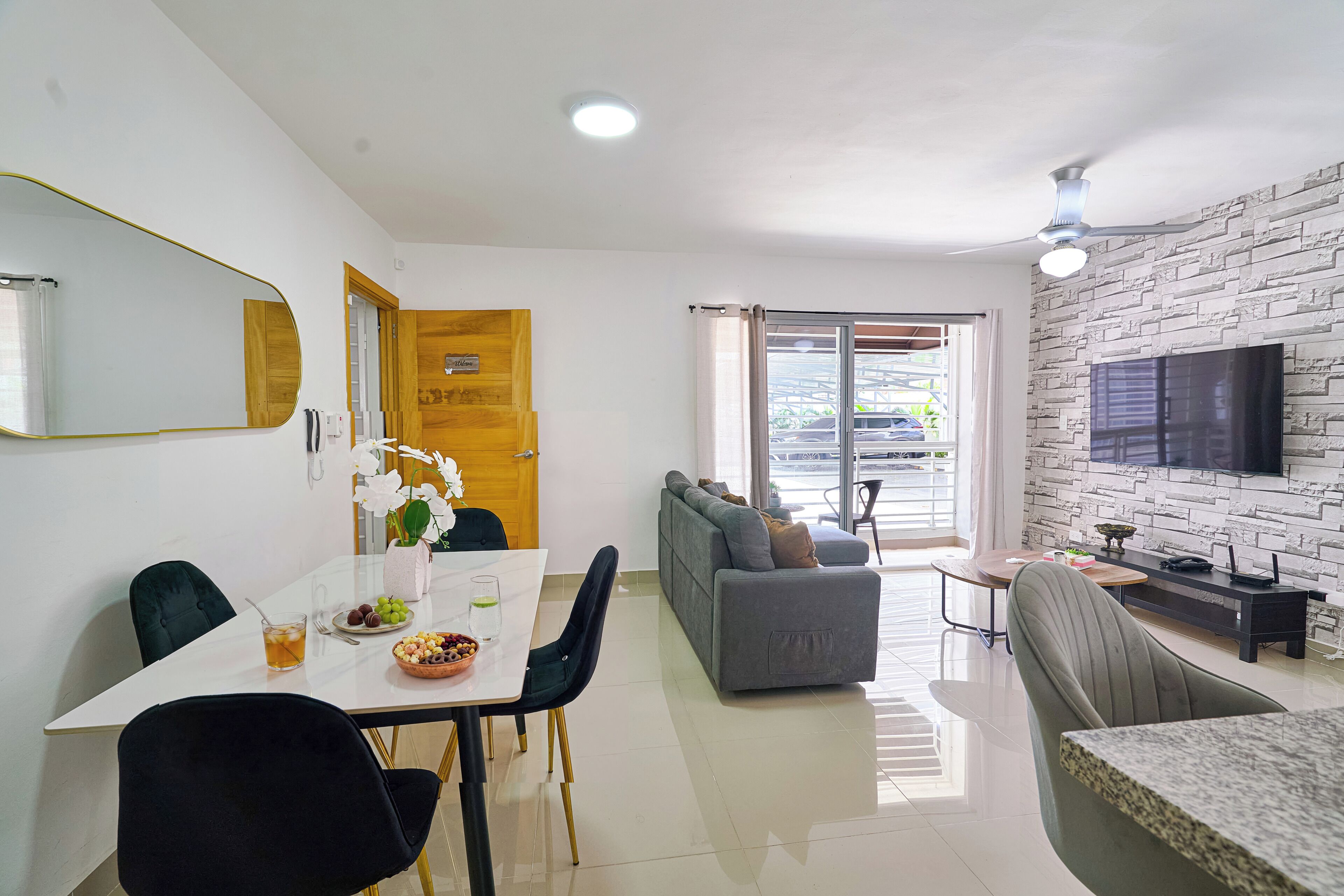 A modern living room with a sofa, dining table, and chairs. There is a television mounted on the wall and a door leading to a balcony.