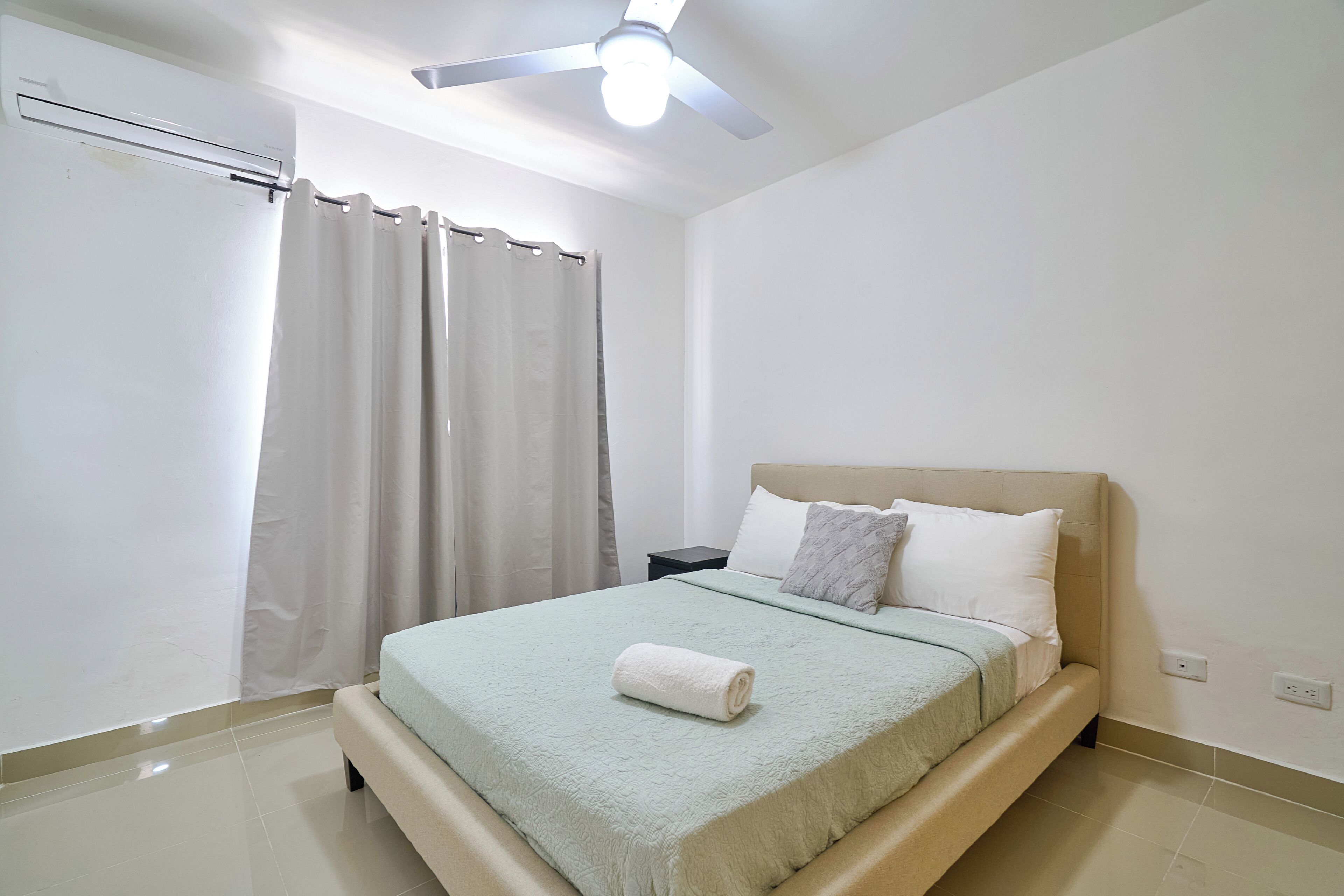 Une chambre à coucher avec un lit, une table de chevet, un ventilateur de plafond et une fenêtre avec des rideaux.
