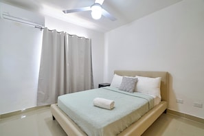 Un dormitorio con cama, mesita de noche, ventilador de techo y ventana con cortinas.