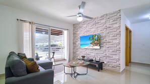 A living room with a grey sofa, a small round table, a ceiling fan, a TV on a stone-textured wall, and a sliding glass door leading to a balcony. - Charming 3-bedroom apartment with Wi-Fi, AC in welcoming Santo Domingo Este. (Santo Domingo Este)