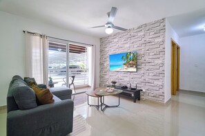 A living room with a grey sofa, a small round table, a ceiling fan, a TV on a stone-textured wall, and a sliding glass door leading to a balcony. - Charming 3-bedroom apartment with Wi-Fi, AC in welcoming Santo Domingo Este. (Santo Domingo Este)
