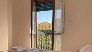Una cama bien hecha con ropa blanca, una ventana que ofrece vistas a un edificio y follaje, y una lámpara en el techo.