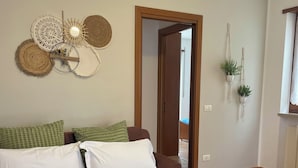 Una cama bien hecha con colcha verde, almohadas blancas y una toalla doblada. Se observan adornos en la pared y una puerta que da a otra habitación.