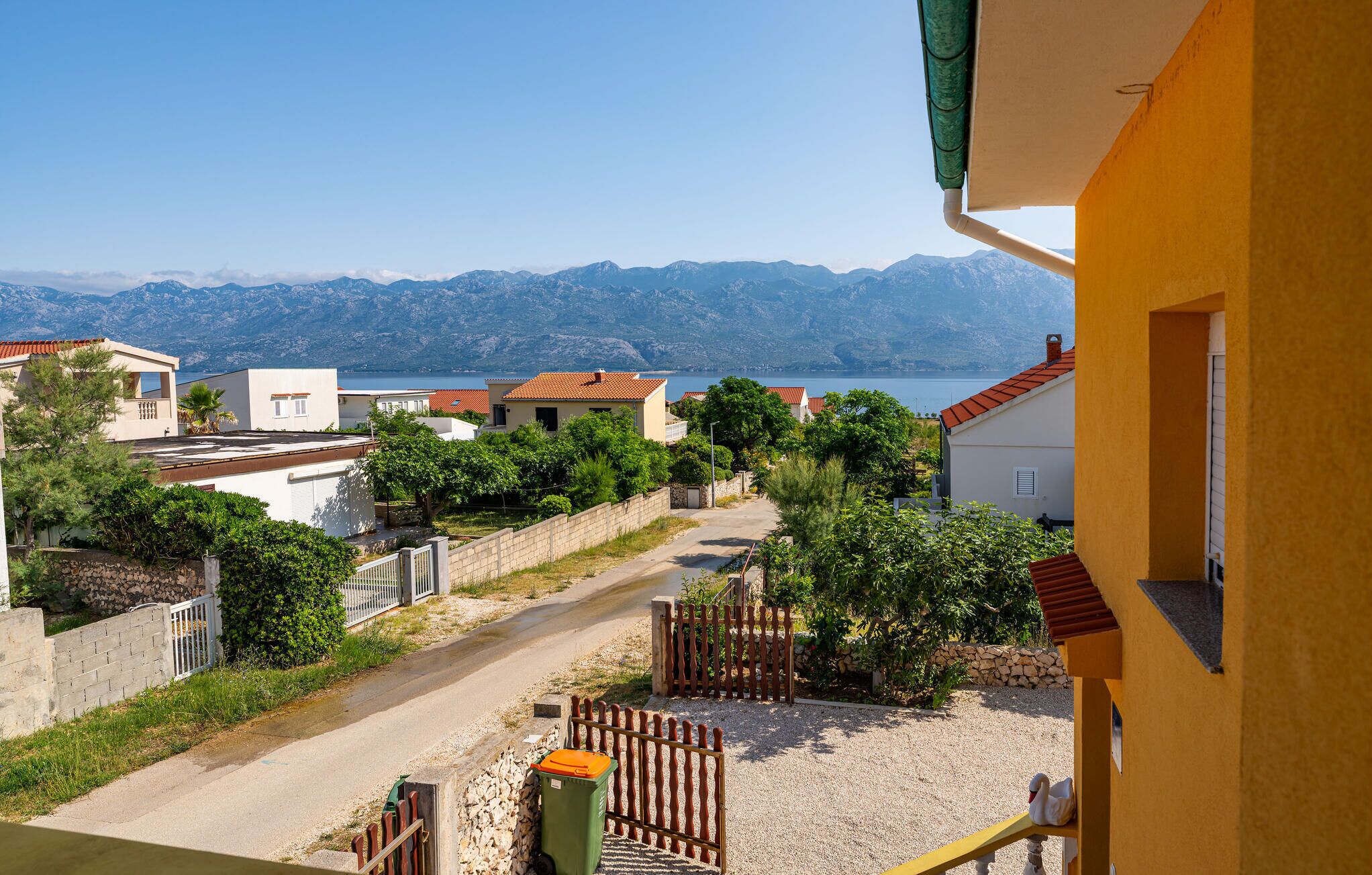A coastal residential area with houses, a clear view of the sea, and a mountain range in the background.