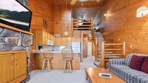 A cozy cabin interior with a stone fireplace, wooden furniture, a kitchen area, and a staircase leading to an upper level. - Scenic 2BR Loft Stay Steps from Pelican Lake (Breezy Point)