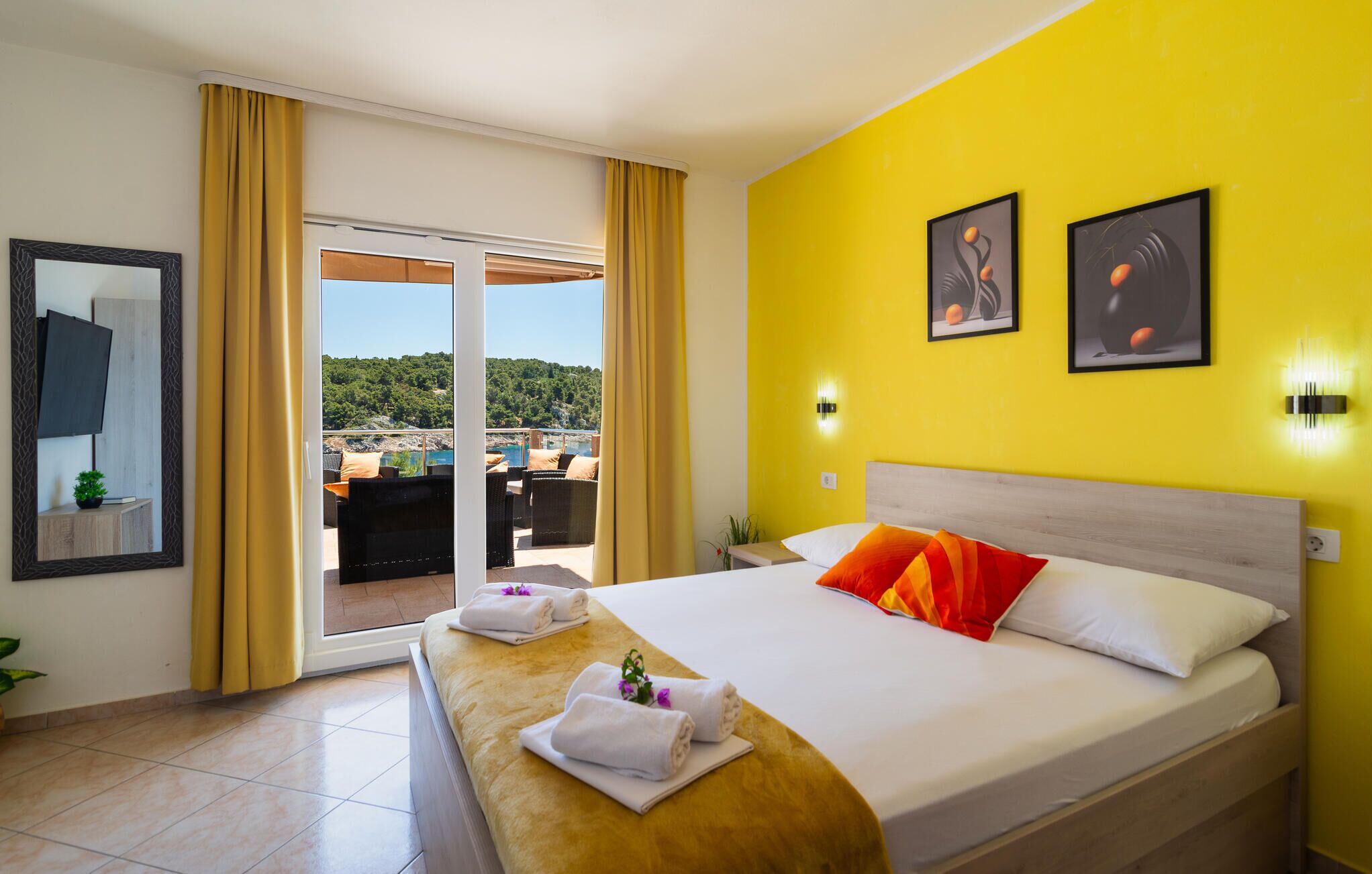 A hotel room with a bed, a wooden side table with towels, a view of a balcony with a scenic landscape, and two wall-mounted artworks.