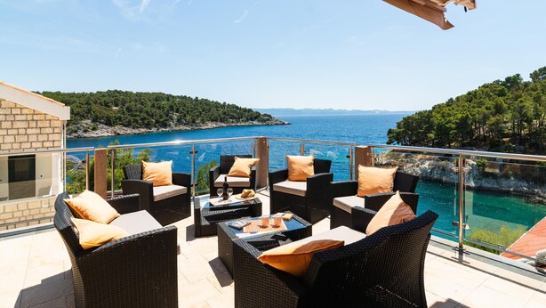 A balcony with wicker furniture, a table with drinks, and a view of the sea and hills.