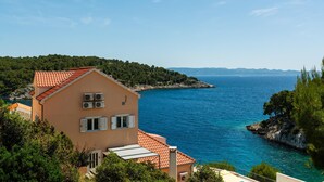 A coastal building with a red-tiled roof, overlooking a clear blue sea and a hilly landscape.