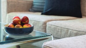 A bowl of fruit on a glass table in a living room with a couch and pillows in the background.