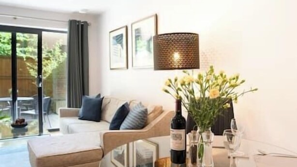 A modern living room with a glass dining table set for two, a sofa, and a large window.