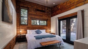 A bedroom with a wooden ceiling, a large bed with a bench, a nightstand, and a window with a view of trees. - Luxury S Austin Home + Casita (Austin)