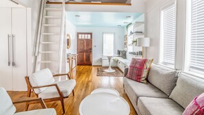 A spacious living room with a wooden floor, a grey sofa, white armchairs, a white coffee table, and a wooden ladder leading to a loft.