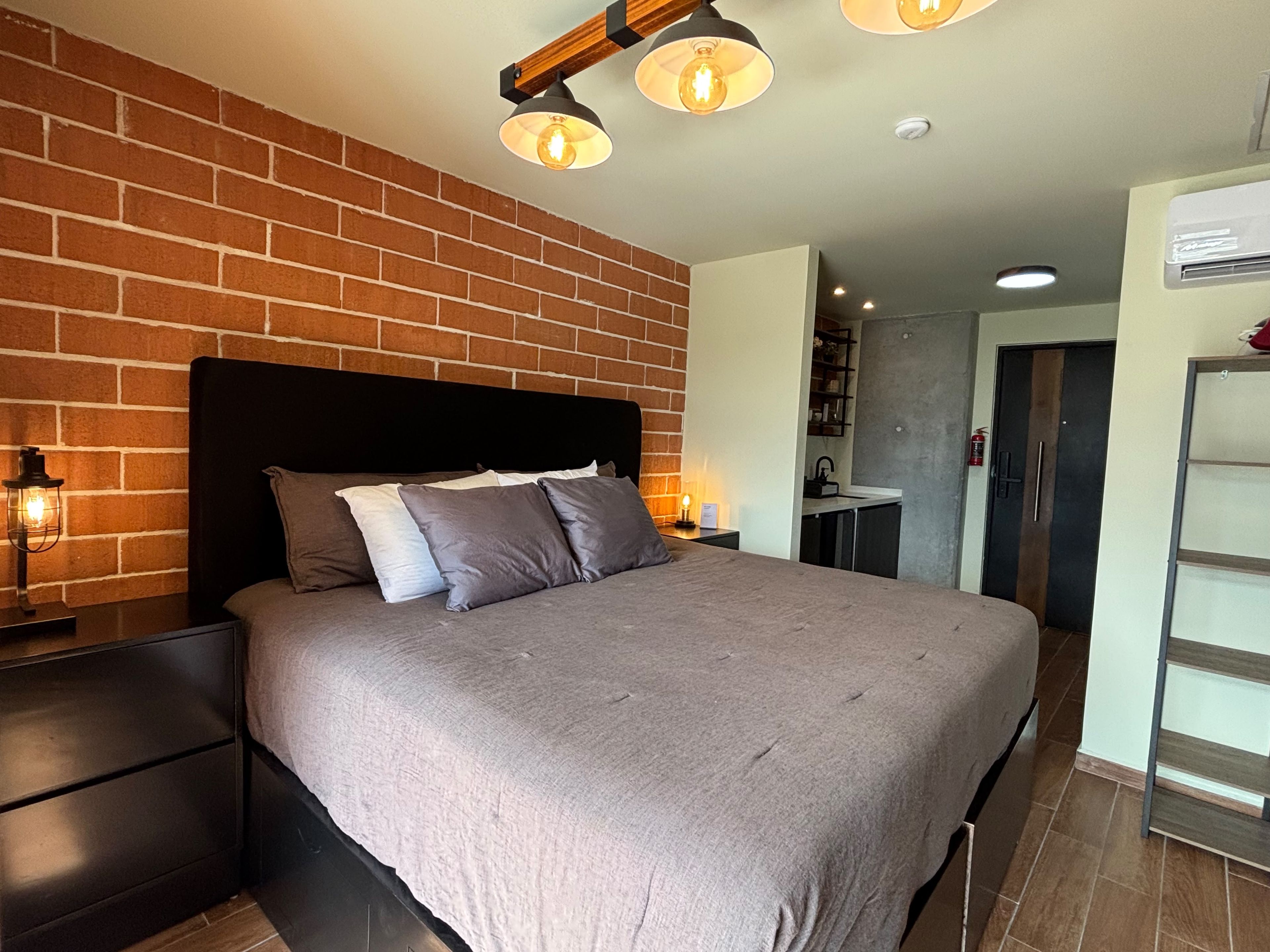 Un dormitorio con pared de ladrillo, cama con ropa de cama gris, mesita de noche con lámpara y lámpara de techo moderna.