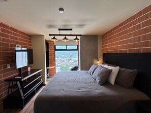Quarto com uma cama grande, televisão presa na parede e vista para a cidade pela janela.