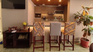 Una cocina moderna con un televisor de pantalla plana, armarios de madera y una isla central. Hay dos taburetes de bar con tapicería estampada y una planta en maceta.