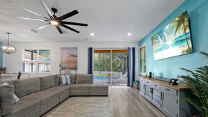 Ein modernes Wohnzimmer mit einer Eckcouch, einem Deckenventilator und einem großen Fenster mit Blick auf einen Pool.