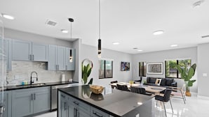 A modern kitchen with grey cabinets, a black countertop, and a central island. There is a dining area with a table and chairs, and a sofa. - Brown House (Miami)