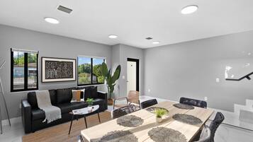 A modern living room with a dining area, featuring a black leather sofa, a wooden dining table with placemats, and a large window with a view of greenery.