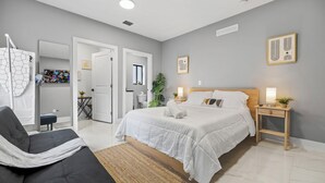 A modern bedroom with a large bed, a bedside table with a lamp, a chair, and a view of a bathroom through an open door. - Brown House (Miami)
