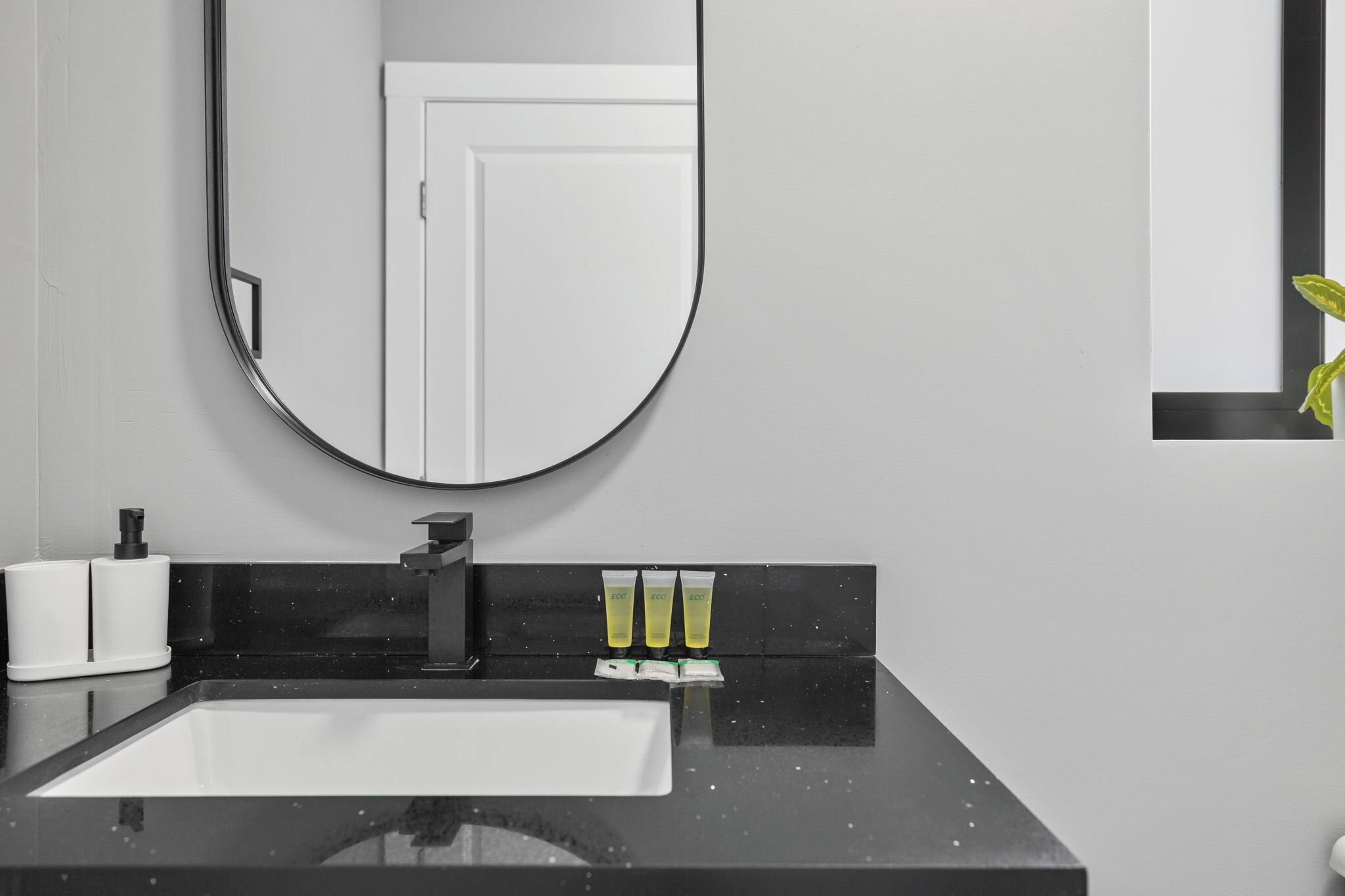 Un baño moderno con encimera negra, lavabo blanco y espejo. Hay tres tubos de loción sobre la encimera.