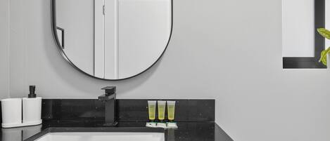 Un baño moderno con encimera negra, lavabo blanco y espejo. Hay tres tubos de loción sobre la encimera.