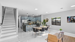 A modern kitchen with a central island, a dining area with a round table, and a staircase leading to another level. - Brown House (Miami)