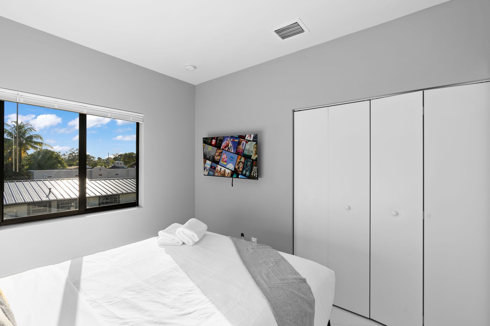 A modern bedroom with a large bed, a wardrobe, a window with a view of palm trees, and a wall-mounted artwork.