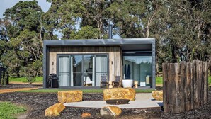 A modern house with a large glass door, a patio with seating, and a wooden fence. - Chalet Coast | Walburra Brook Off-grid Luxury stay (Metricup)