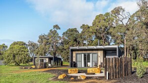 Dos cabañas modernas con grandes puertas correderas de vidrio, rodeadas por una cerca de madera y un jardín ajardinado con rocas y un árbol pequeño.