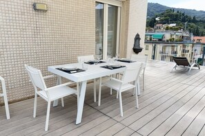 A terrace with a white table set for dining, white chairs, and a view of a cityscape with buildings and hills.