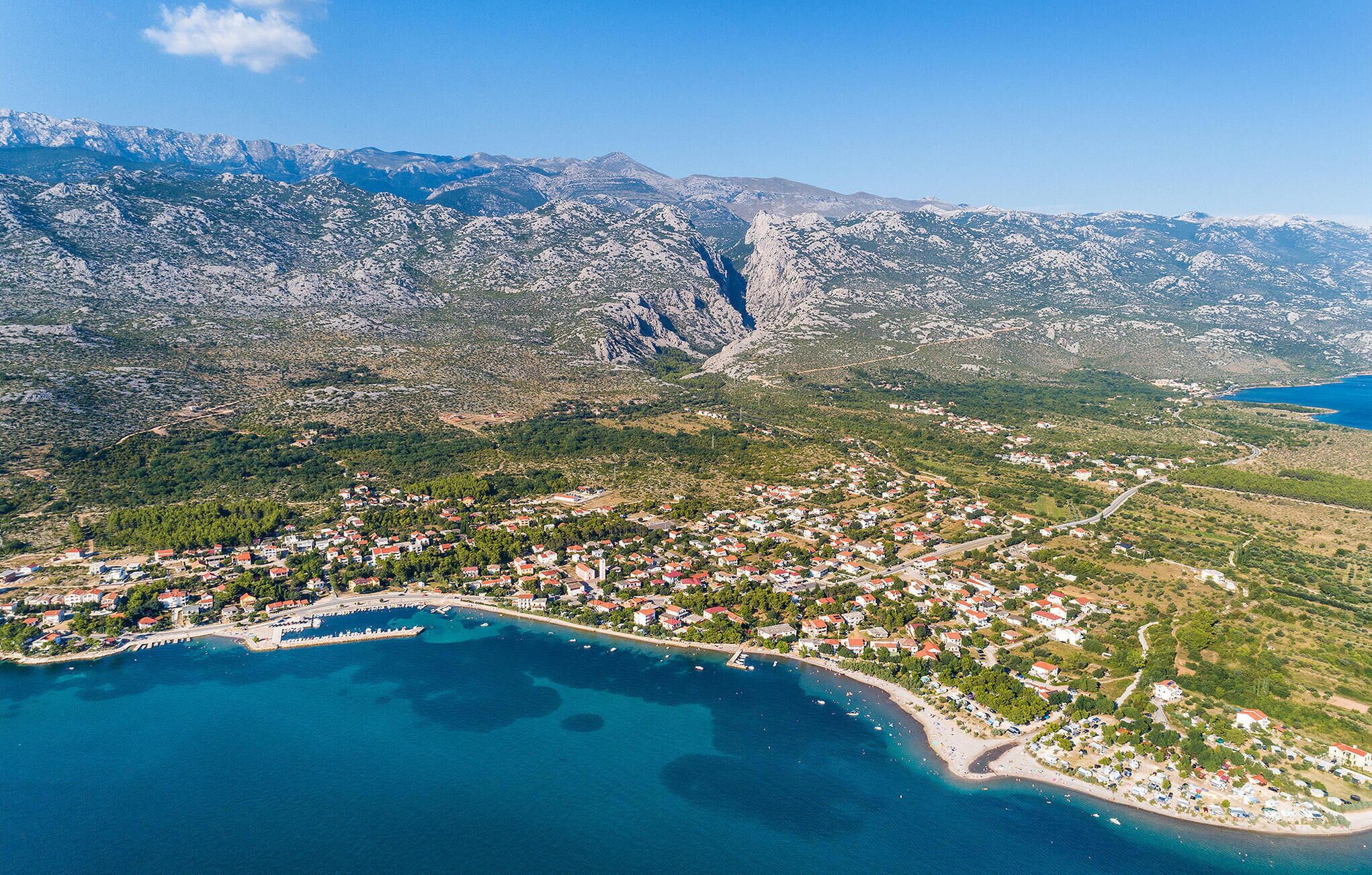 Une vue aérienne d’une ville côtière, avec une chaîne de montagnes en arrière-plan, où l’on aperçoit un mélange de bâtiments résidentiels et d’espaces verts.