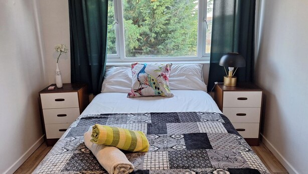Una cama bien hecha con una colcha estampada, dos almohadas, una lámpara de noche y un pequeño florero con flores sobre una mesita de noche.