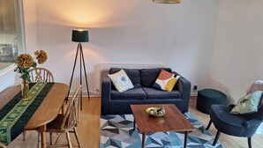 Un salón con un sofá azul oscuro, una mesa de comedor de madera, una lámpara de pie y una mesita auxiliar con un cuenco.
