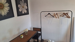 Una habitación con una mesa de madera, una silla, un pequeño jarrón con flores y un perchero con perchas y zapatos.