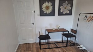 Una habitación con piso de madera, una puerta blanca, una pequeña mesa con dos sillas y dos flores enmarcadas en la pared.