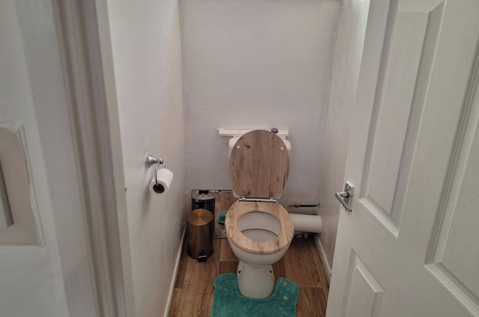 A small, narrow bathroom with a wooden toilet, a white wall, and a green mat on the floor.