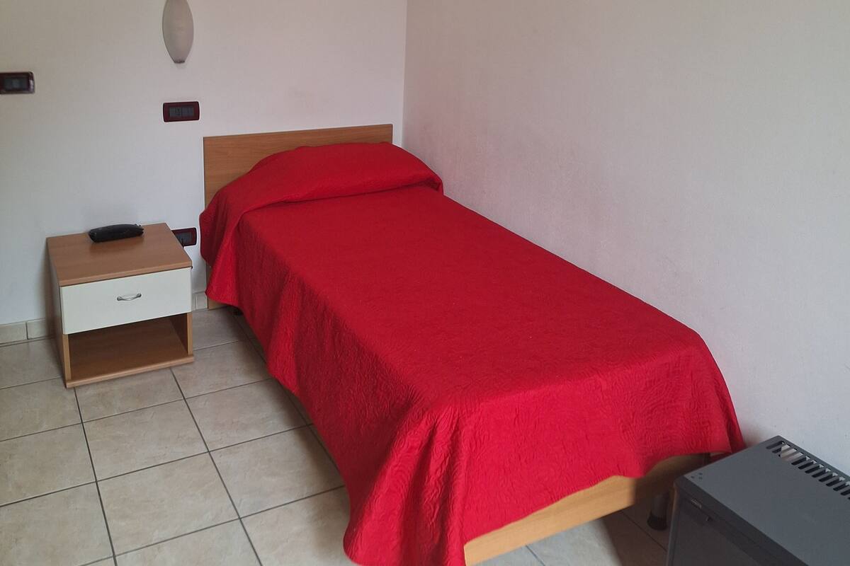 A single bed with a red bedspread, a wooden nightstand with a drawer, a small table with a black object on top, and a gray appliance on the floor.