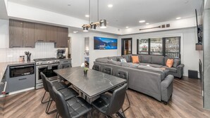 A modern kitchen with a dining area, a large gray sectional sofa, and a TV mounted on the wall. - Designed Specifically for Large Families (Canmore)