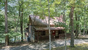 A rustic cabin with a porch, surrounded by dense forest. - Cabin on the lake! Hot tub, fishing, pool access! (Sevierville)