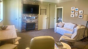 A living room with a stone fireplace, a flat-screen TV mounted above it, a white sofa, a coffee table, and a dining table with chairs. - Absollut 9 - Central location (Mount Hotham)