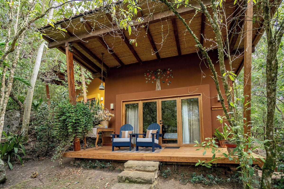 A wooden deck with two chairs and a table, surrounded by lush greenery and a thatched roof structure.