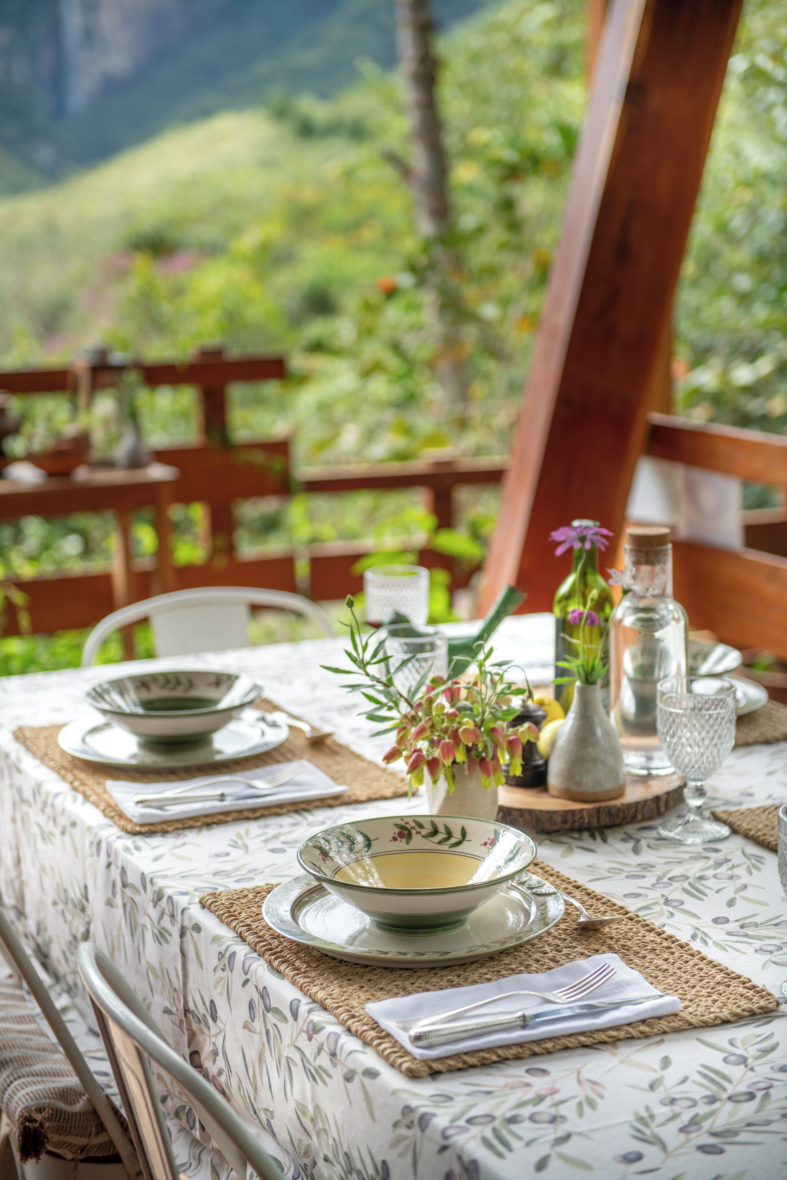 Et spisebord dækket med tallerkener, skåle, bestik og en vase med blomster, med grøn vegetation i baggrunden.