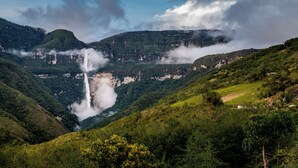 Paisagem montanhosa com cachoeira, vegetação exuberante e céu limpo.