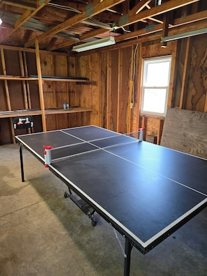 Una mesa de ping pong en una habitación con paredes de madera, una ventana y una estantería con objetos.