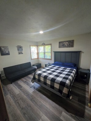 A bedroom with a bed, a sofa, a nightstand, a window with blinds, and a wall-mounted picture.