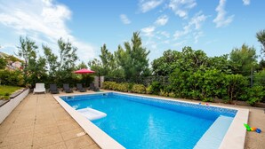 Una piscina rectangular con borde de azulejos, rodeada por un patio empedrado. Se observan sillas de descanso y una sombrilla roja al fondo.