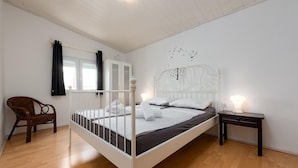 Un dormitorio con estructura de cama de metal blanco, una silla de madera, una mesita de noche con lámpara y una ventana con cortinas.