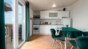 Cocina compacta con armarios blancos, microondas, estufa y pileta. Hay una mesa de comedor con sillas verdes y una pequeña planta en maceta.